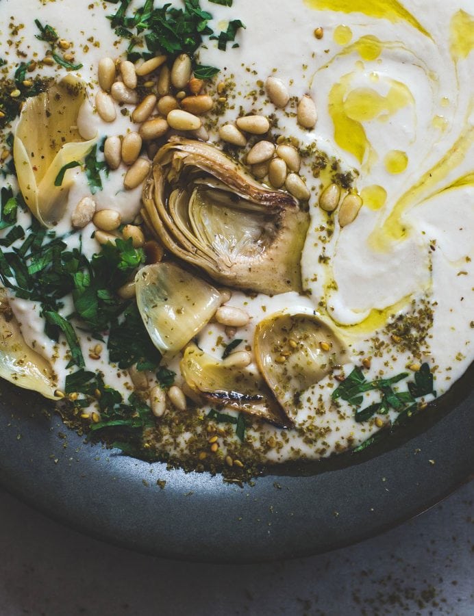 white bean hummus in a black bowl, topped with artichokes and pine nuts, olive oil, and parsley