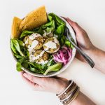 Bowl of salad topped with baked falafel, held in someone's hands, overhead angle.