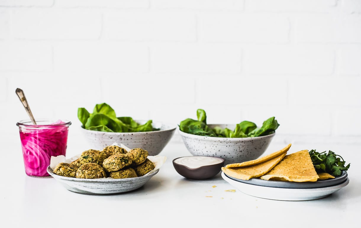 two bowls of salad in backround, with bowl of baked falafel and socca at forefront, next to jar with pickled red onions. eye level photo.