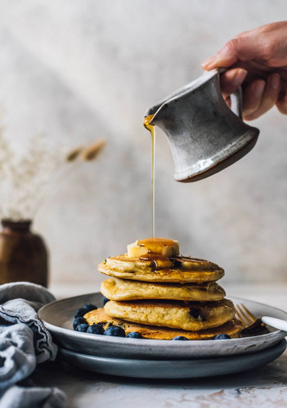 pouring maple syrup over a stack of blueberry pancakes