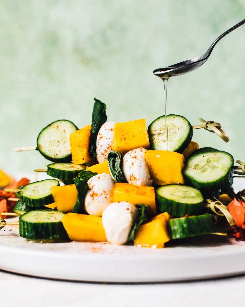 drizzling white balsamic vinegar over cucumber mango skewers stacked on a white plate