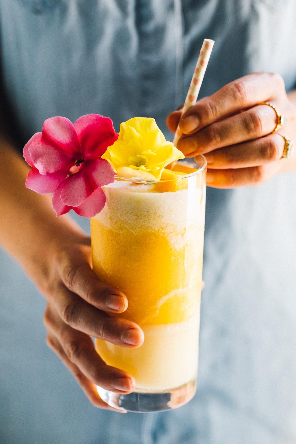 girl holding a mango lava flow cocktail, with one yellow and one pink flower as a garnish.