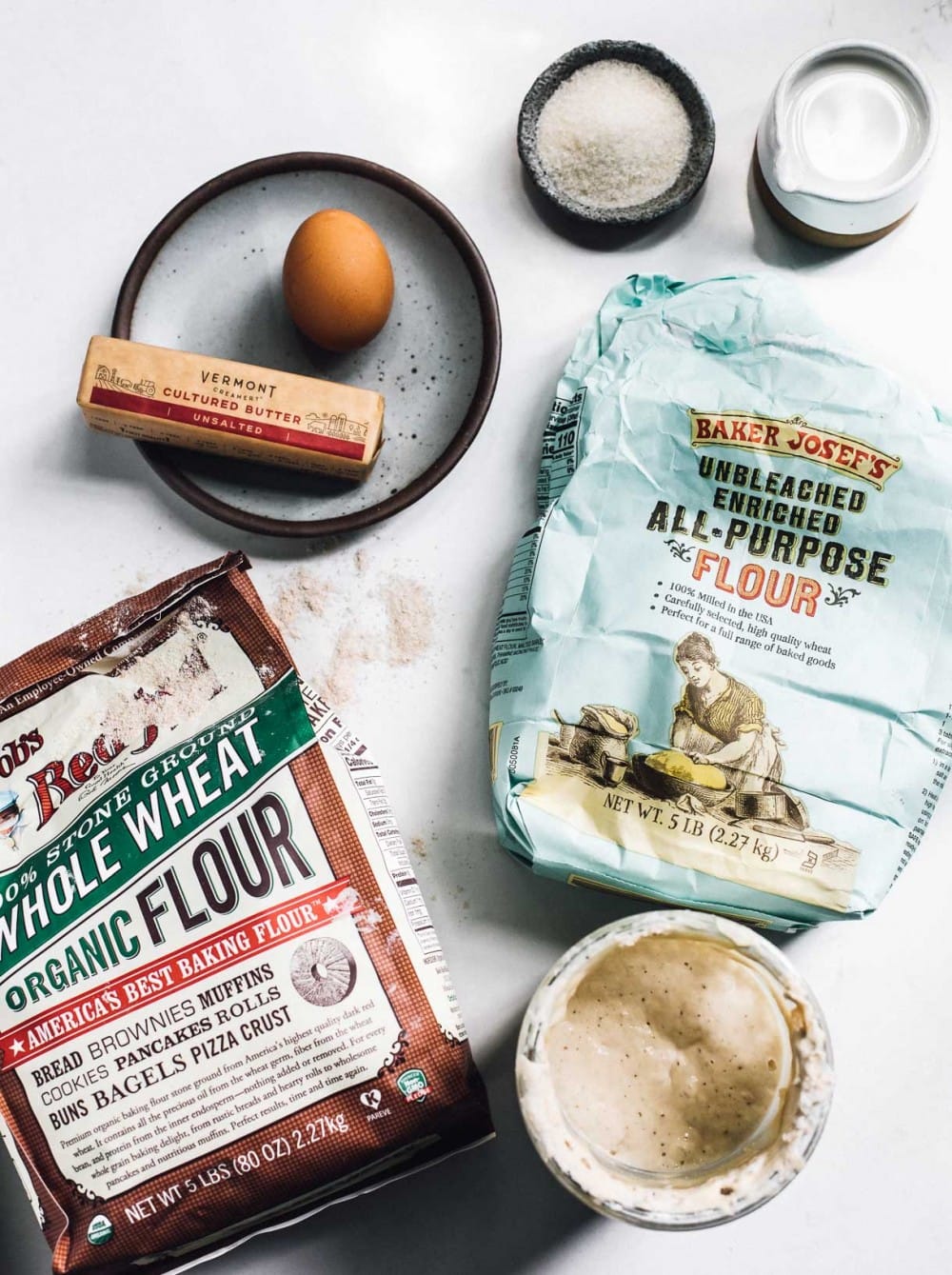 a bag of whole wheat flour, all purpose flour, a stick of butter, one brown egg, sugar, and milk, and sourdough starter, laid out on a kitchen counter. 