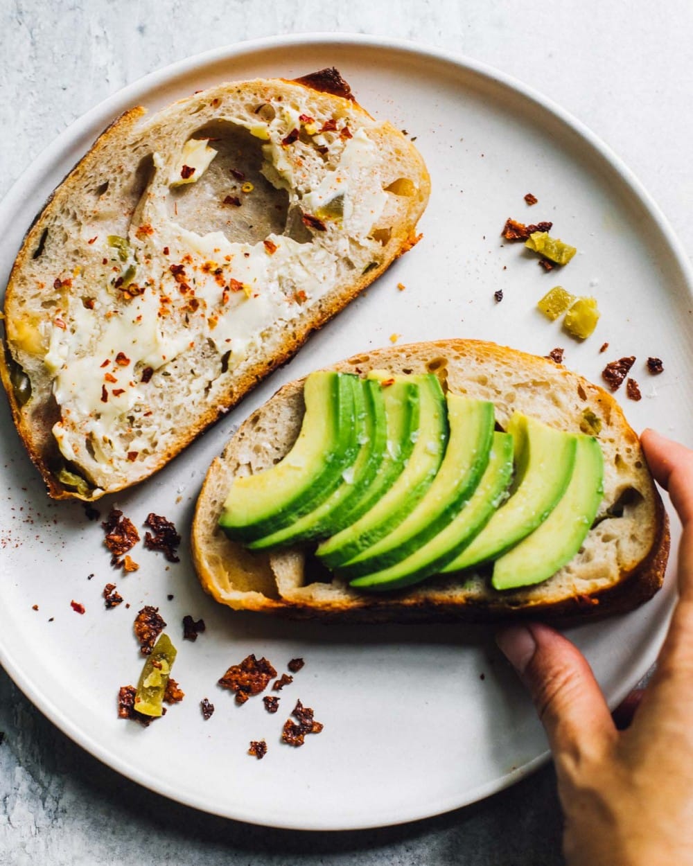 two slices of sourdough bread on a white plate, one with avocado, the other with butter and red pepper flakes.