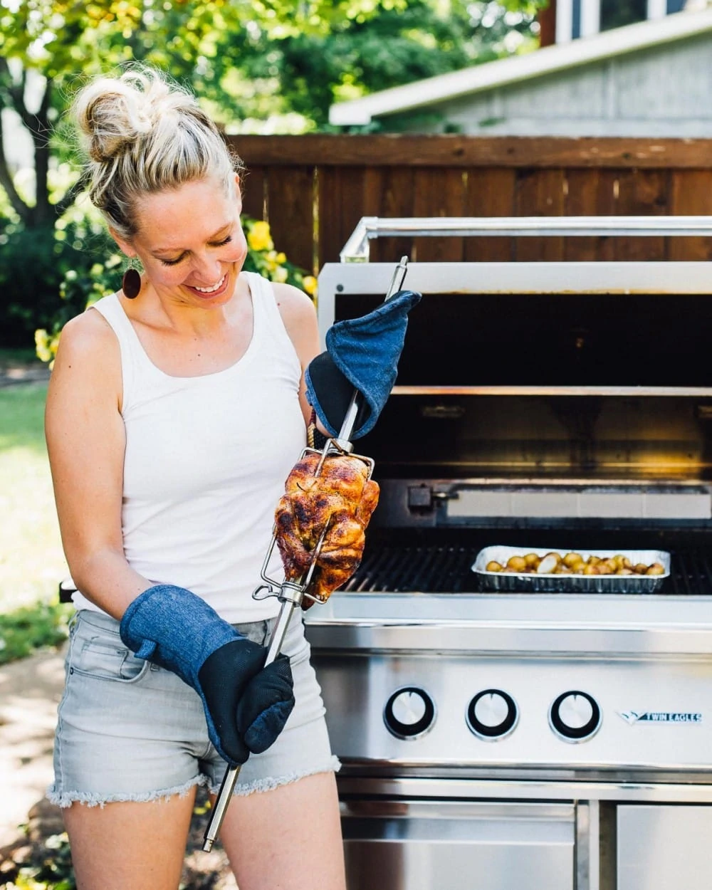 girl holding a rotisserie chicken attached to a spit, open grill in backround