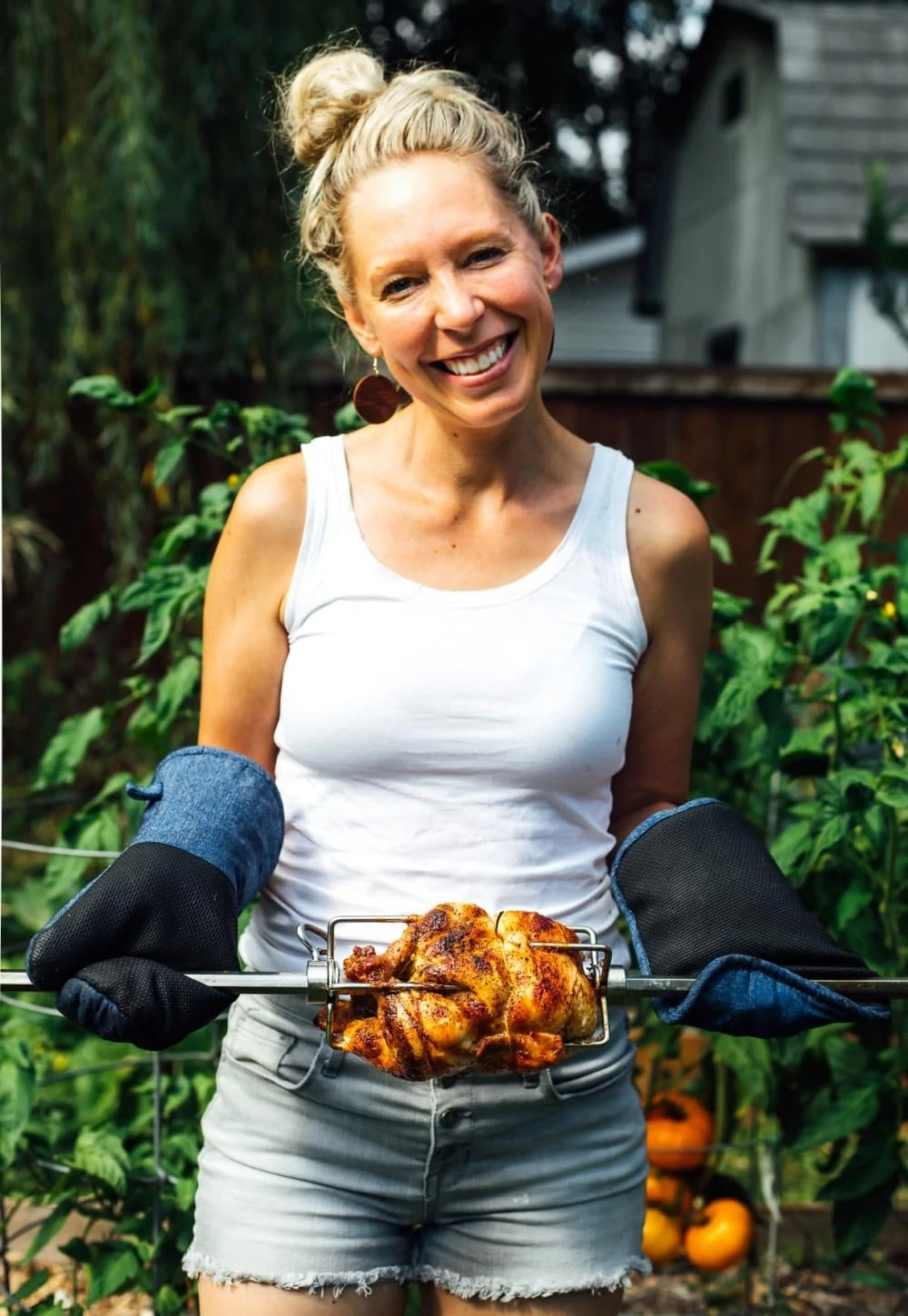 girl holding rotisserie chicken in front of garden. she is wearing white tanktop and grey shorts