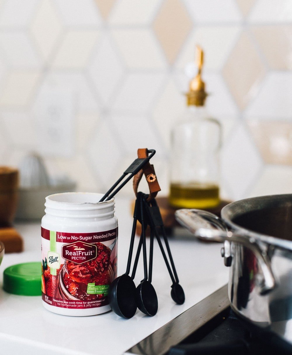 a opened container of Ball Fruit Pectin with a set of measuring spoons peeking out of it. bottle of olive oil in the backround.