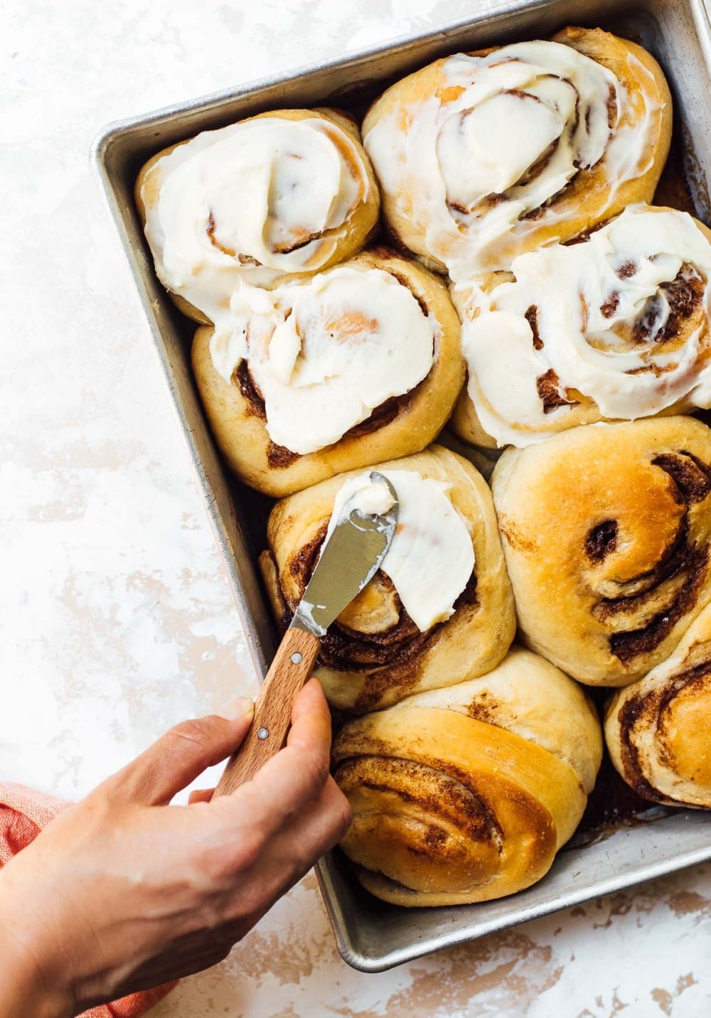 spreading cream cheese frosting on a pan of cinnamon rolls