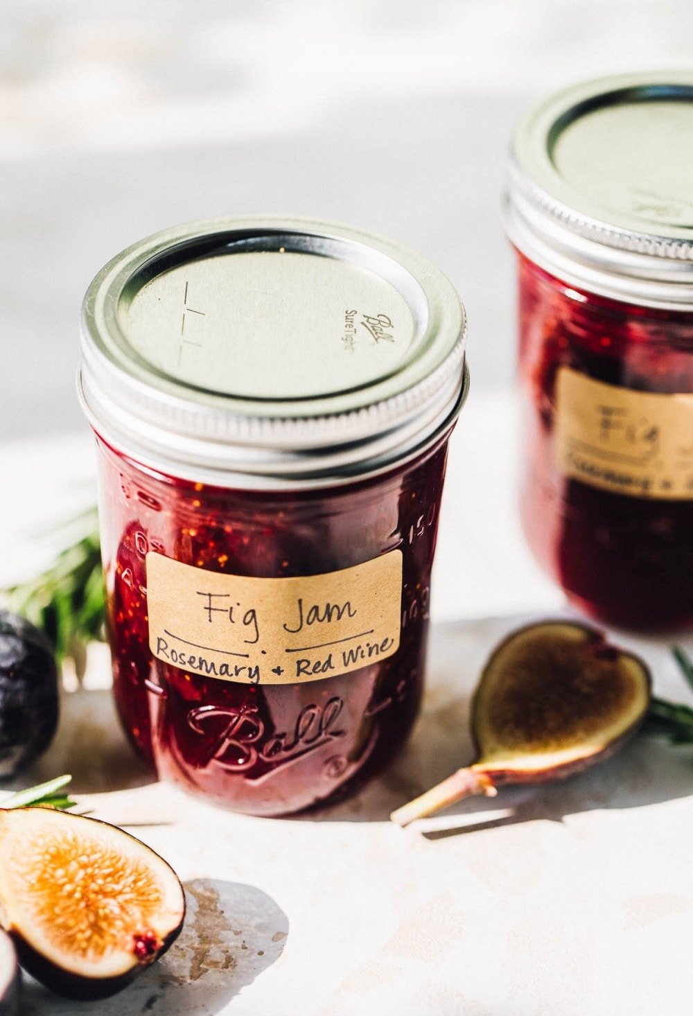 two jars of fig jam, unopened, with a fig and rosemary next to the jars