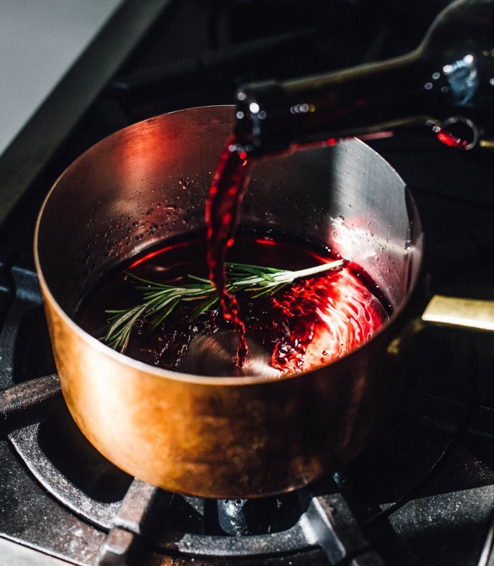 red wine being poured into copper kettle, rosemary also in the pan