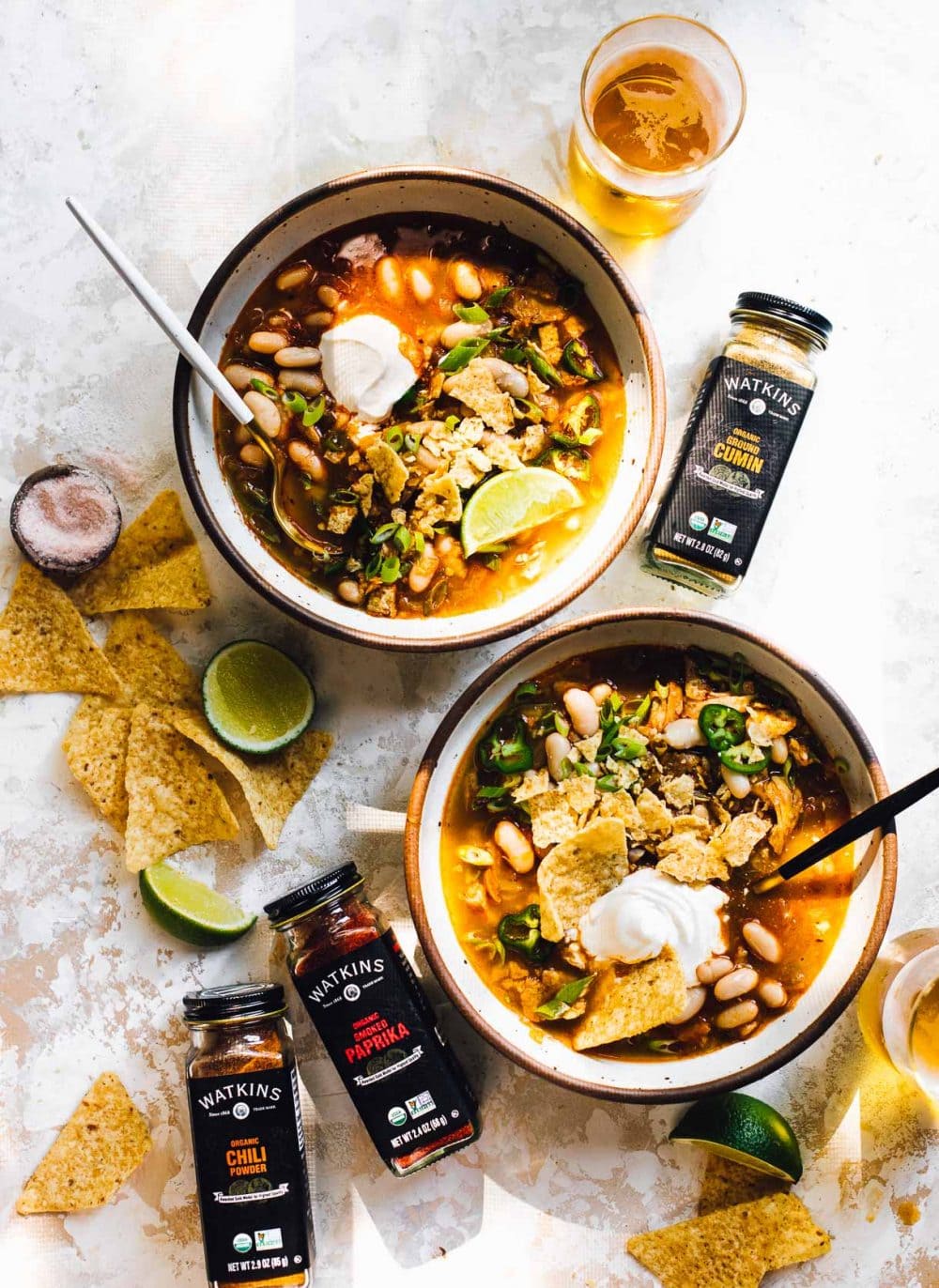 two bowls of green chili chicken soup with tortilla chips scattered around bowls of jars of spices, glass of beer in upper right