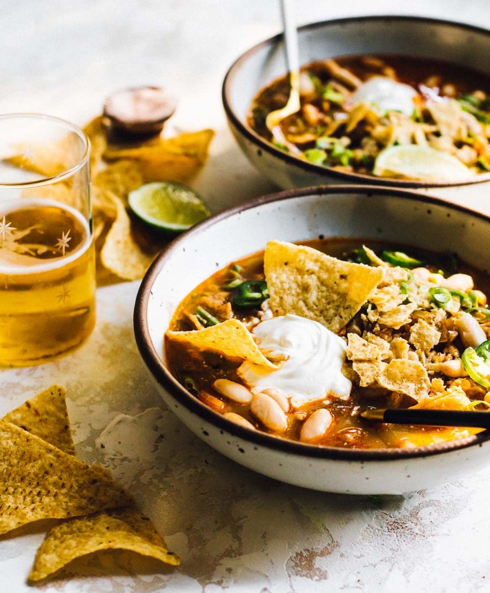 bowl of green chili chicken soup with tortilla chips in it, dollop of sour cream on top, tortilla chips scattered around bowl and glass of beer to the left