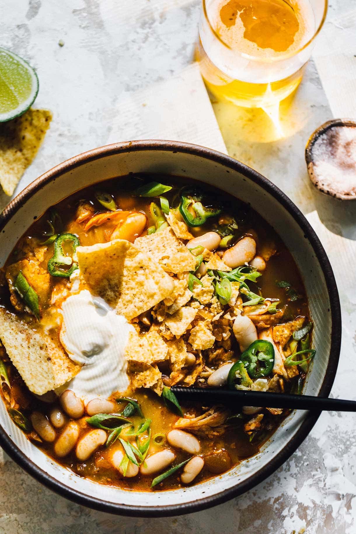 bowl of chicken chili with glass of beer alongside of it, chips crushed on top, as well as dollop of sour cream and green onions.