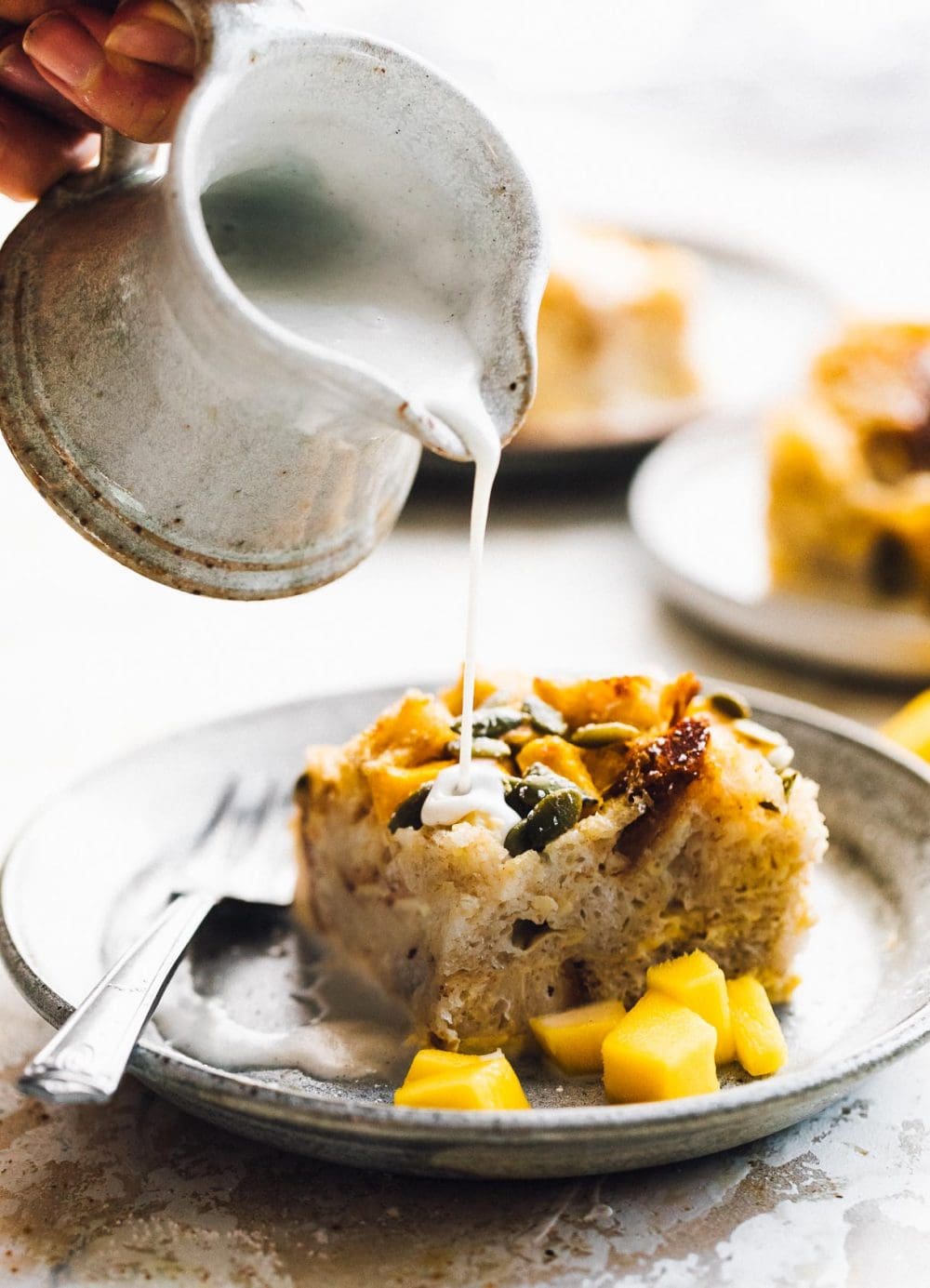 pouring vanilla cream onto bread pudding, with mangos on the side of plate