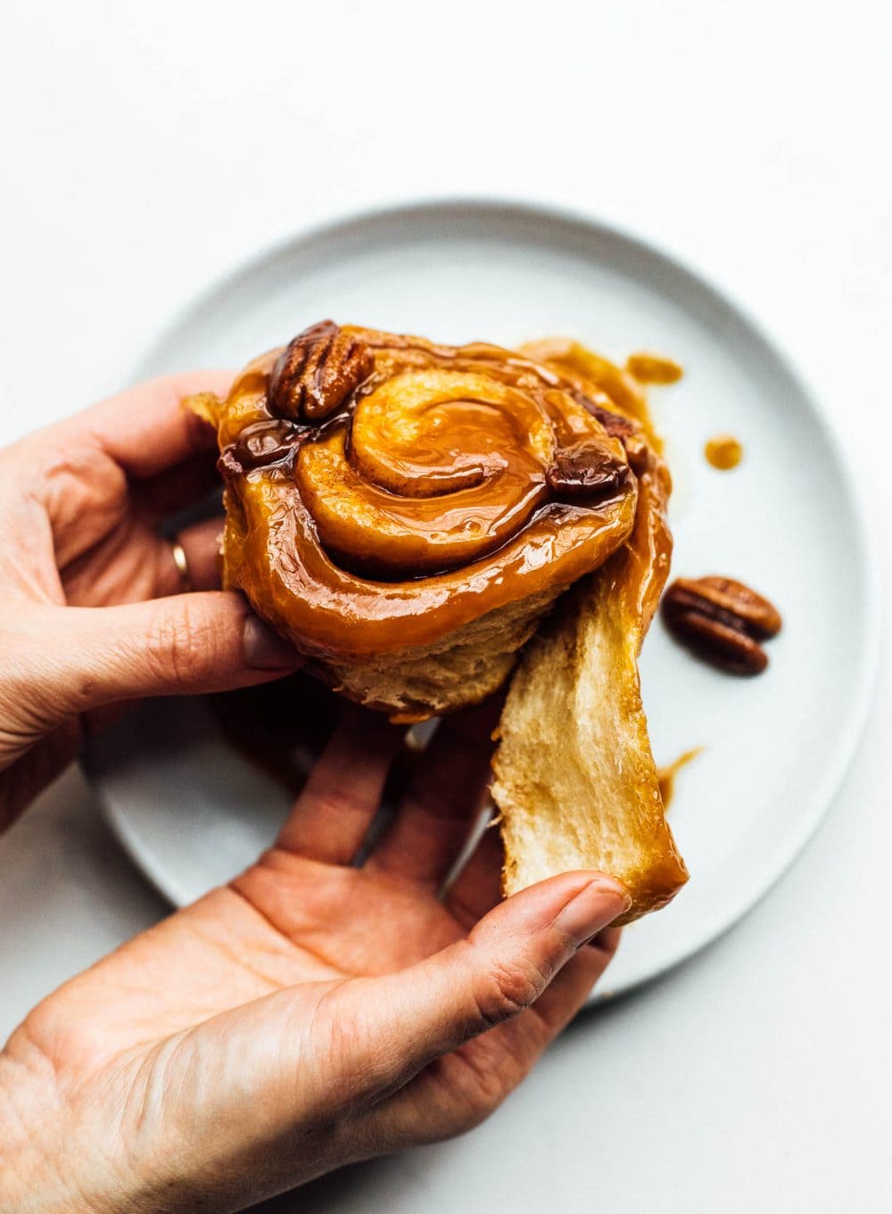 Woman is holding sourdough sticky bun in her hands, tearing some of the dough off. Blue plate is below her hands. 