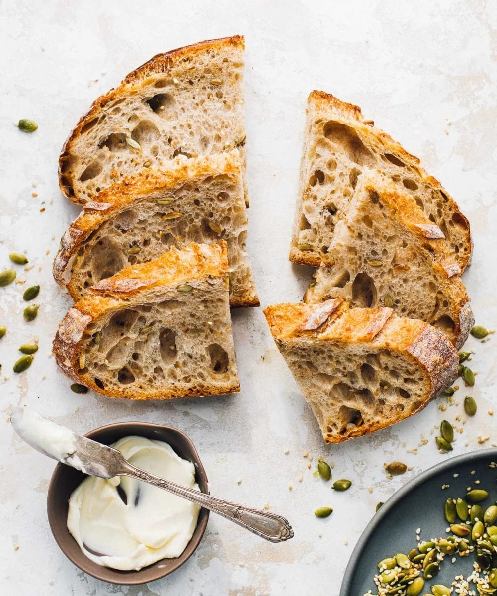 seeded sourdough bread slices cut in half, with a bowl of butter to it's bottom left, and on the bottom right a blue plate with toasted pepita seeds