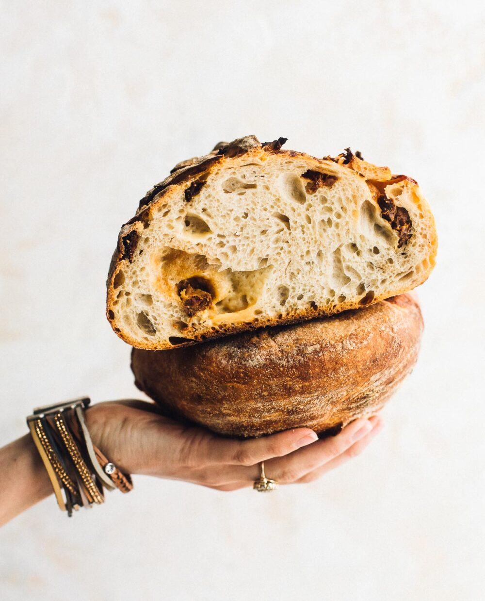 holding a loaf of sundried tomato and cheddar sourdough bread, cut in halves, and the halves stacked on top of one another. woman is wearing a bracelet on her hand. 