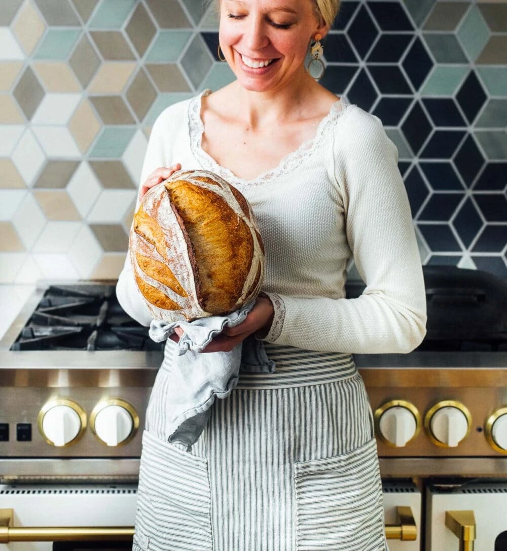 woman holding sourdough bread in front of her oven, she has a white shirt on. 