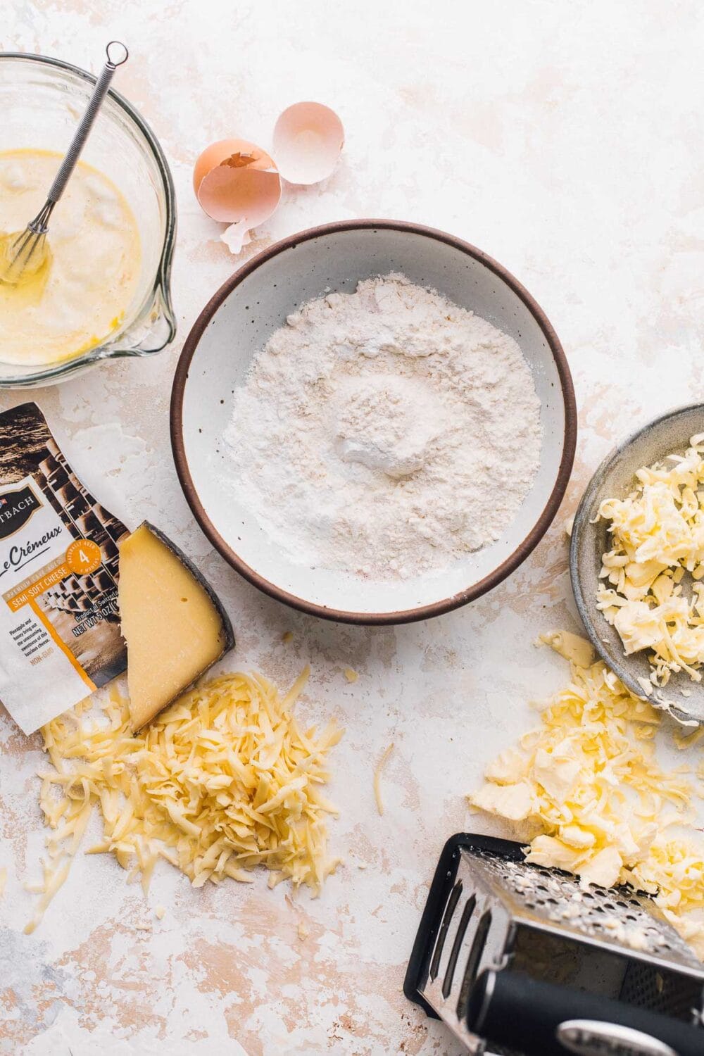 ingredients for sourdough discard biscuits laid out on a white surface, has a bowl of flour in the middle with grated cheese and butter to its side