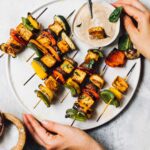 grilled veggie kabobs with dipping sauce on a white plate, woman holding the plate.