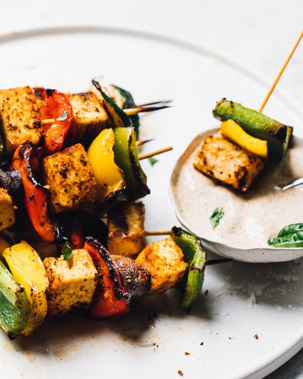 grilled veggie skewers with tofu on a white plate, with side of dipping sauce to the right of plate.