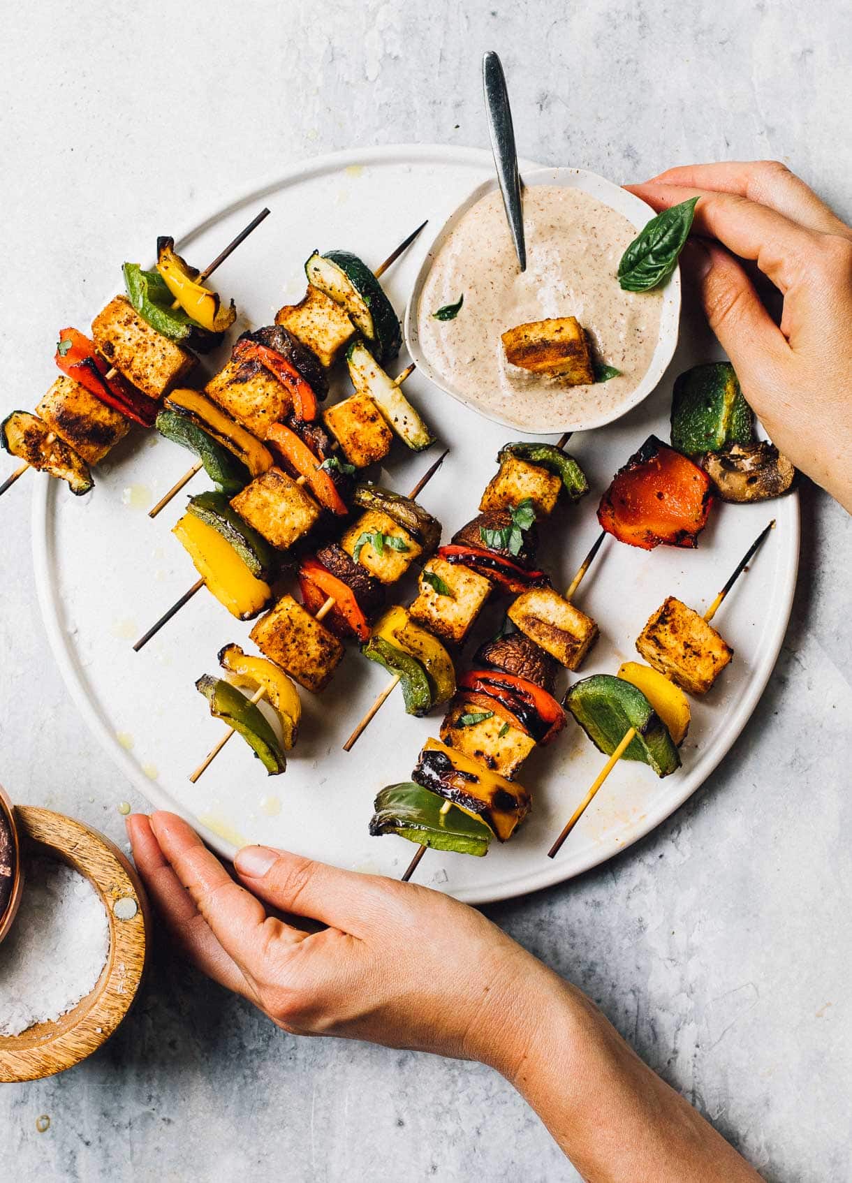 grilled veggie kabobs with dipping sauce on a white plate, woman holding the plate.