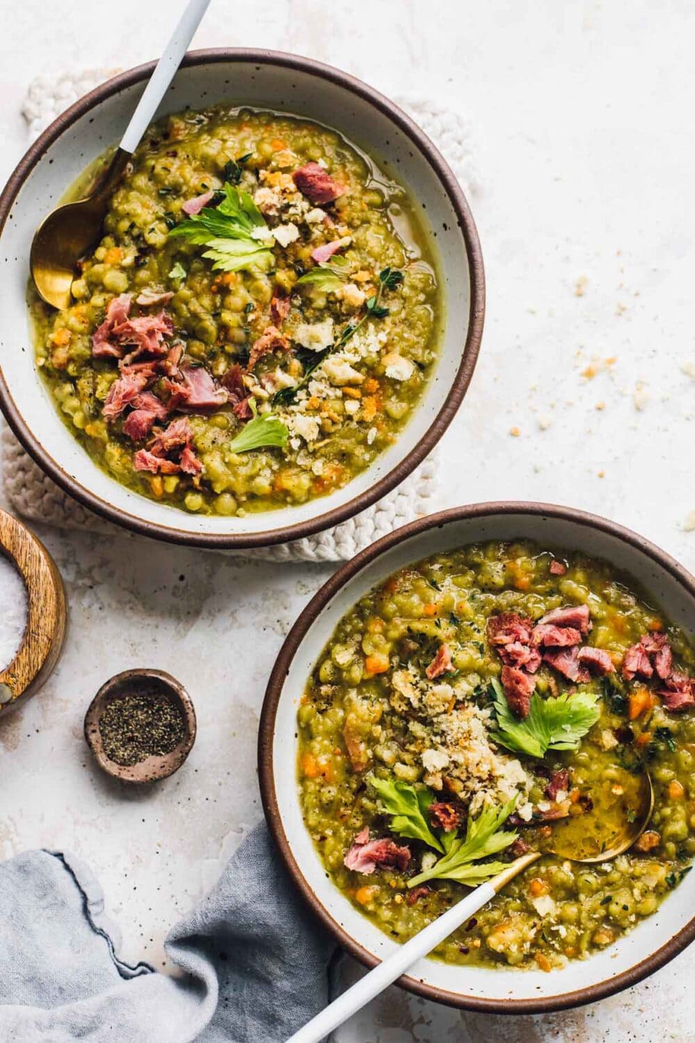 two bowls of split pea soup, with gold spoons, blue linen to the left and salt + pepper.