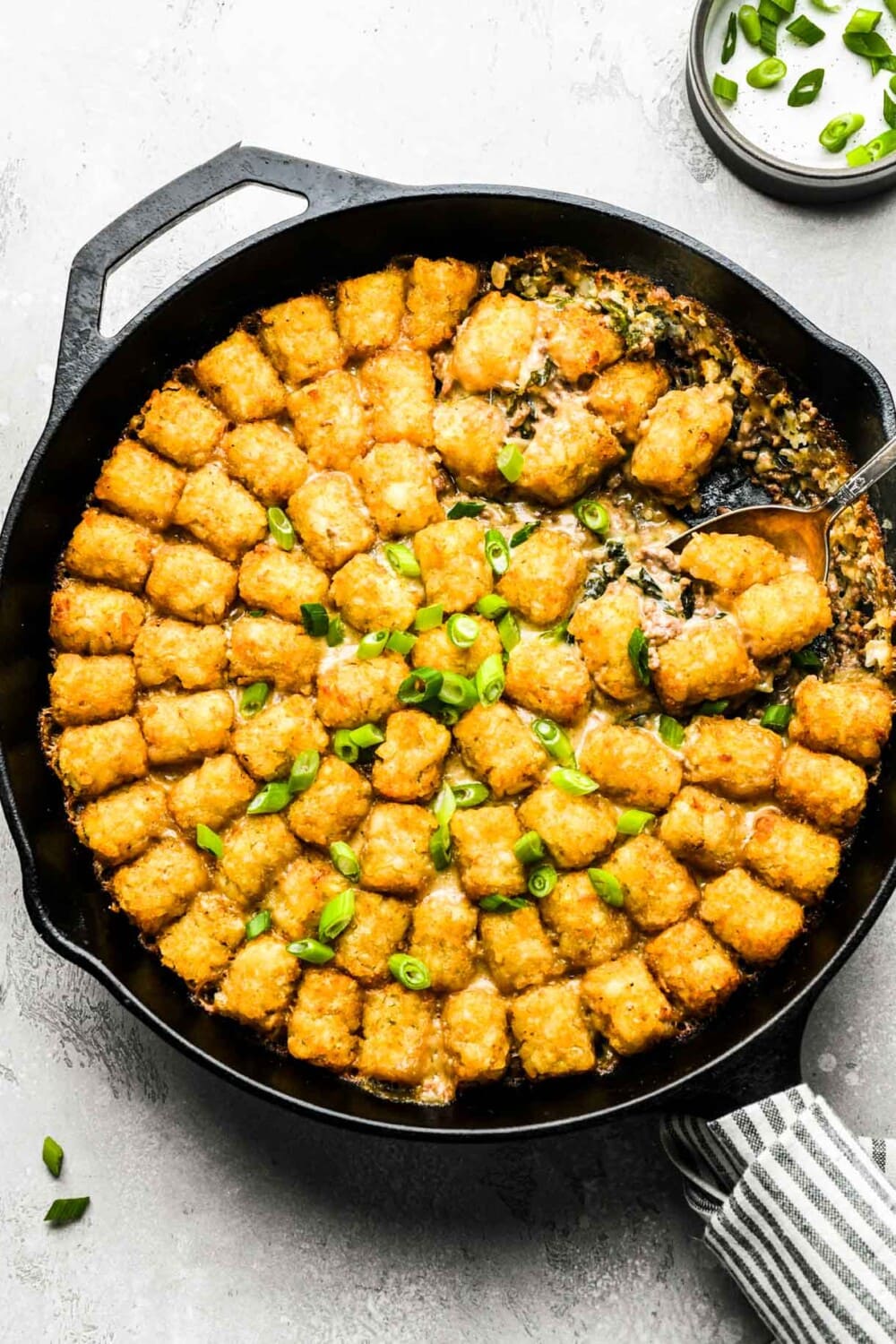 tater tot hotdish in a cast iron skillet with a scoop out of it. linen wrapped around handle.