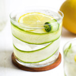 cucumber lemon water in a clear glass, sitting on a wooden coaster