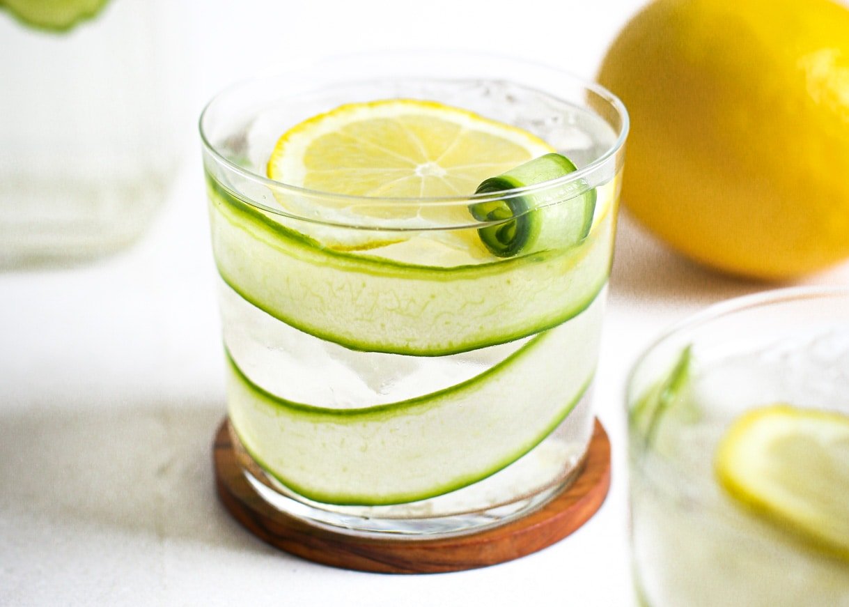 cucumber lemon water in a clear glass, sitting on a wooden coaster