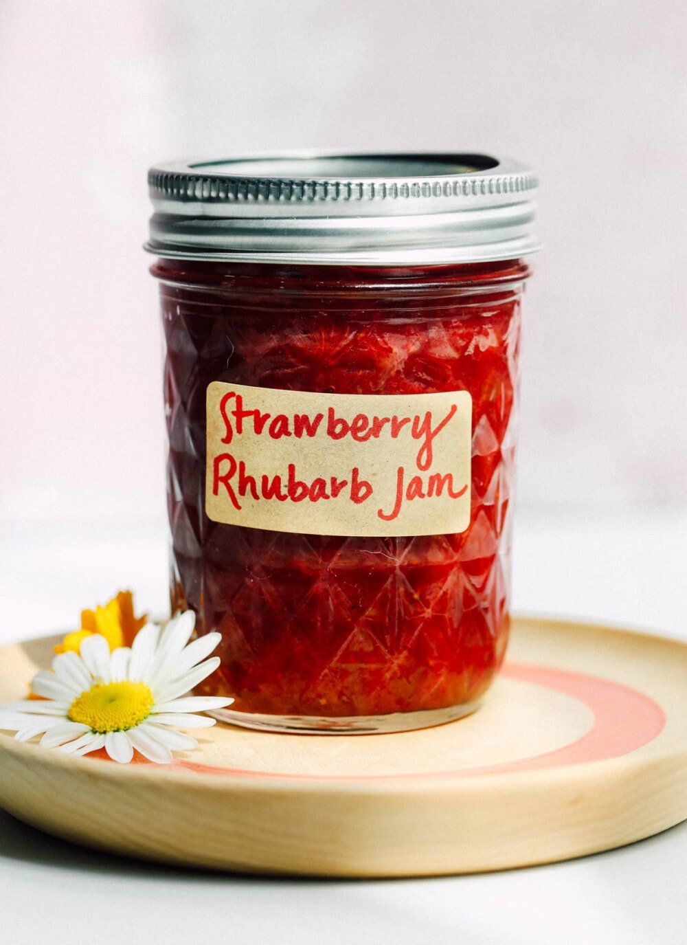 strawberry rhubarb jam in a glass jar with label, sitting on a wooden coaster