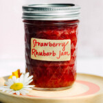 strawberry rhubarb jam in a glass jar with label, sitting on a wooden coaster