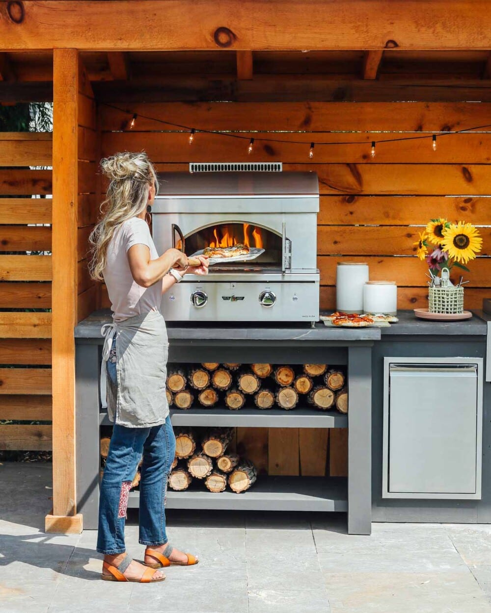 blond woman putting pizza into an outdoor pizza oven