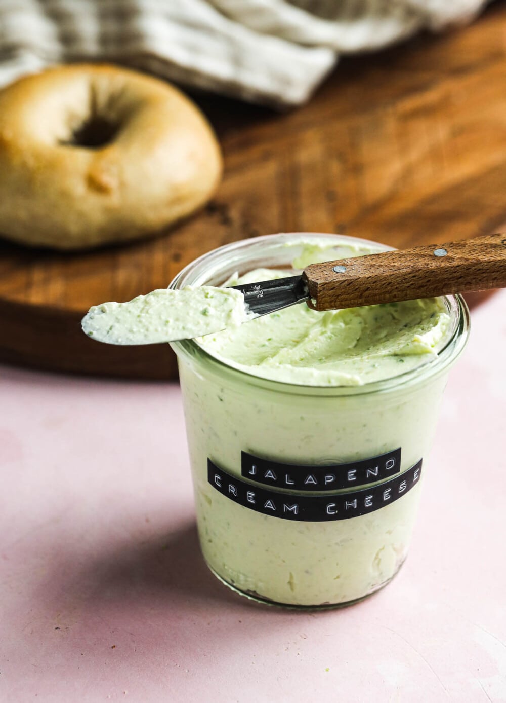 jalapeno cream cheese in a glass jar with knife laying across the top and bagel in the backround