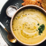 Bowl with green tomato soup on blue plate, with bowl of salt and sourdough bread.