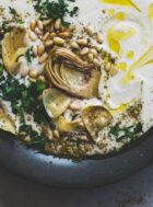 white bean hummus in a black bowl, topped with artichokes and pine nuts, olive oil, and parsley
